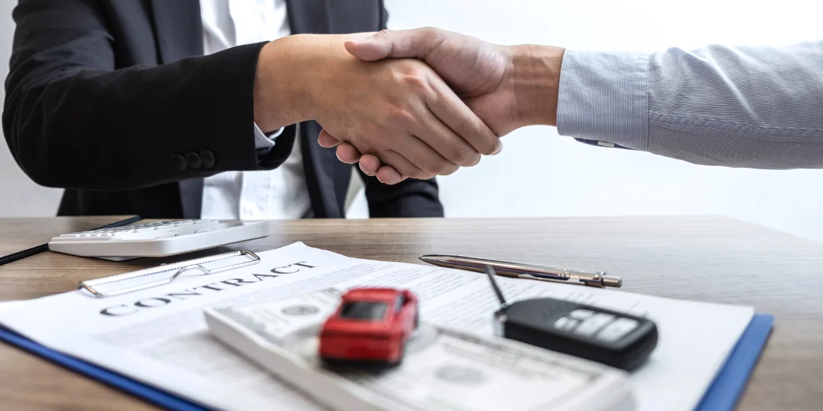 Imagem com duas pessoas dando aperto de mãos com um carrinho embaixo e um contrato representando seguro auto mensal banco do brasil.