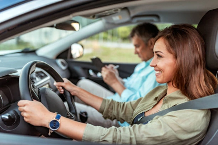 Mulher sorridente dirige veículo, enquanto homem sentado no banco do carona faz anotações. Falando sobre "seguro auto para mulher é mais barato".
