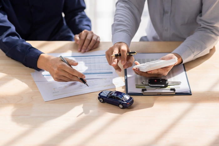 Corretor e cliente analisam apólice de seguro. Na mesa há papéis, uma miniatura de carro e outros itens. Falando sobre a bllu seguro por assinatura.
