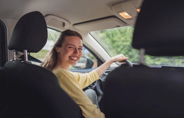 Fato tirada dos bancos traseiros de veículo dirigido por mulher. Ela olha para trás e sorri para a foto. Falando sobre seguro auto mulher.