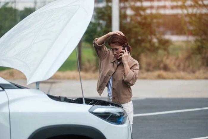Condutora coloca uma das mãos na cabeça, enquanto atende o telefone com a outra. Ela está olhando dentro do capô do seu carro, que está levantado. Falando sobre o melhor seguro de carro para não ficar na mão.