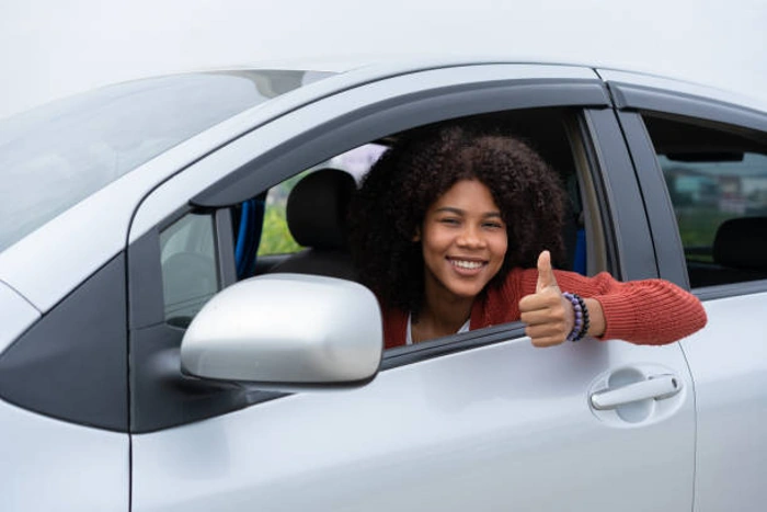 Mulher faz sinal de positivo e sorri de dentro do seu veículo. Falando sobre "seguro auto para mulher é mais barato".