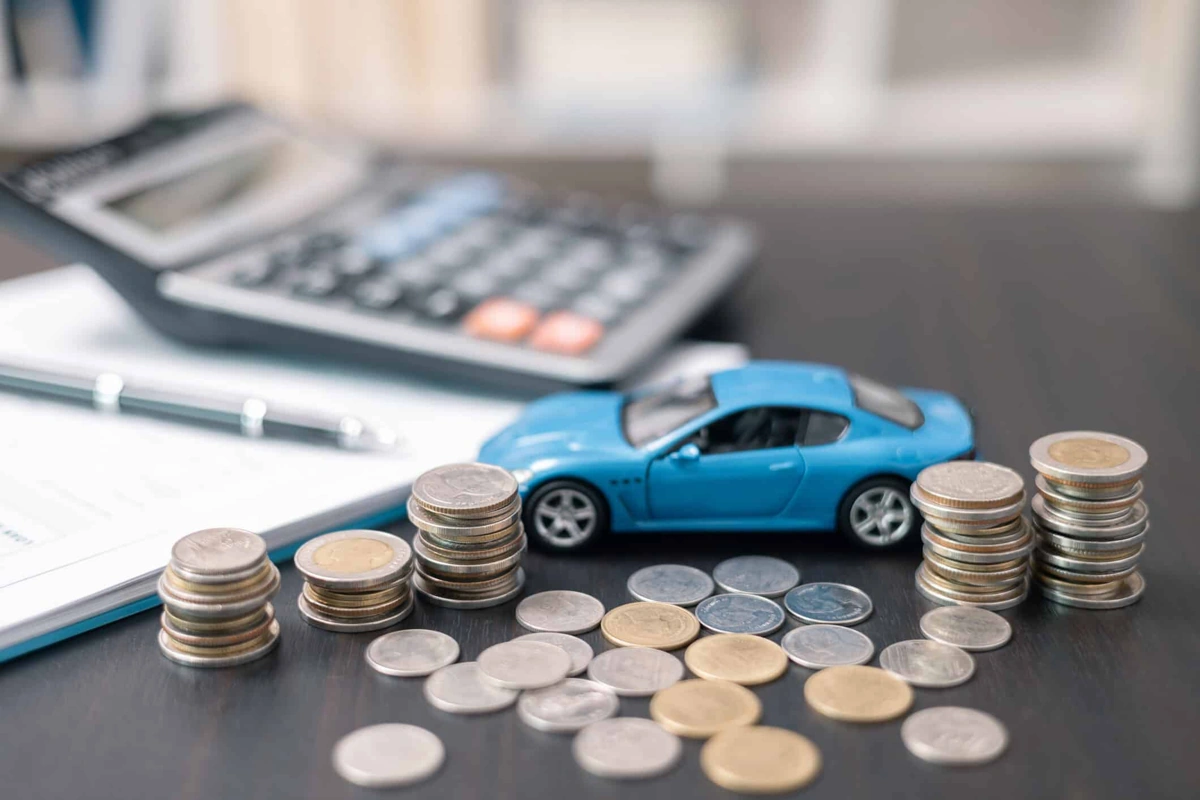 Carrinho de miniatura, na cor azul, sobre moedas. Há ainda na mesa um papel branco, uma caneta e uma calculadora. Falando sobre qual seguradora mais barata.