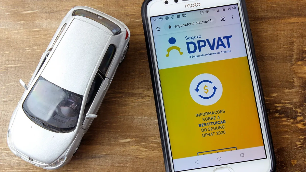 Celular com logomarca do seguro DPVAT na tela, ao lado de uma miniatura de carro na cor cinza. Falando sobre seguro obrigatório para carro.