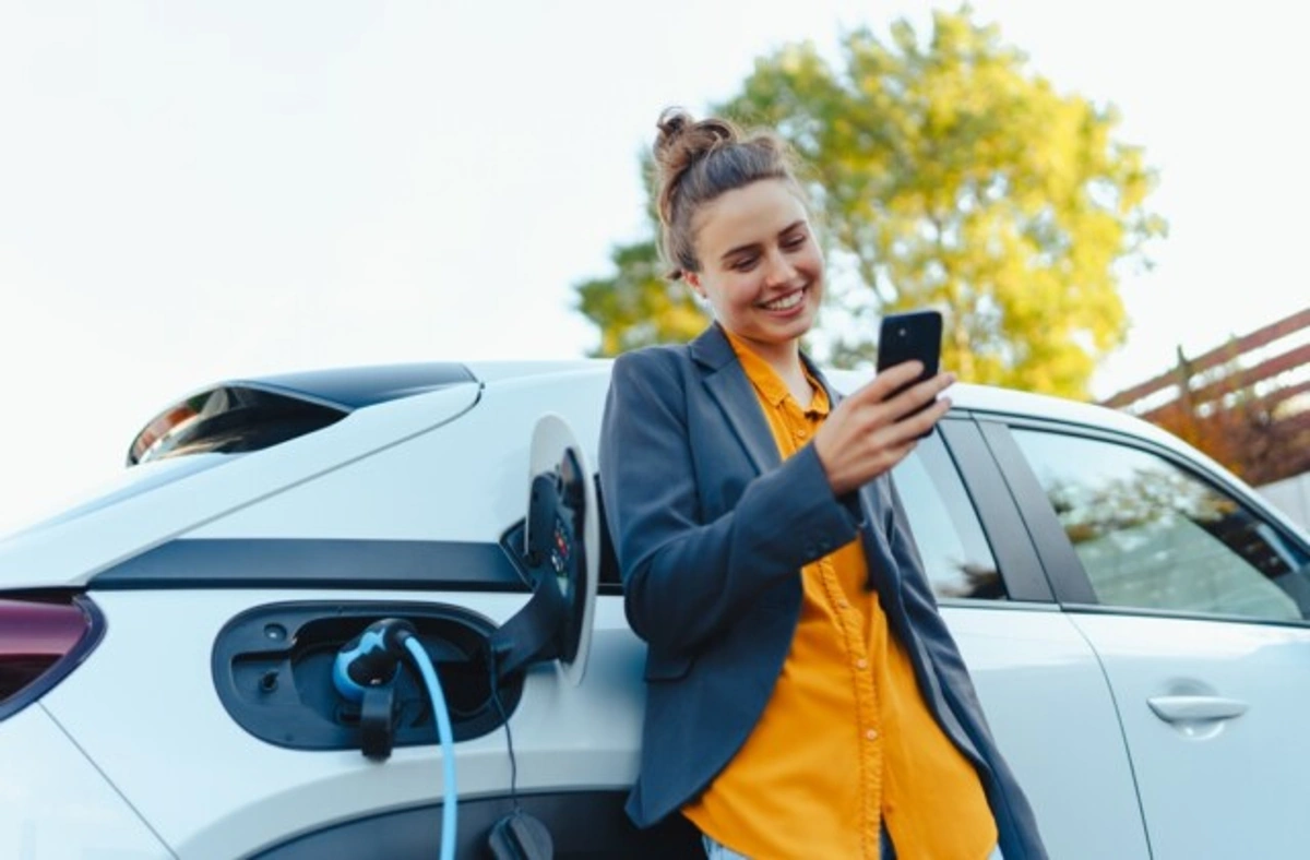 Mulher sorridente mexe no celular enquanto seu carro elétrico carrega. Falando sobre seguro para carro eletrico.