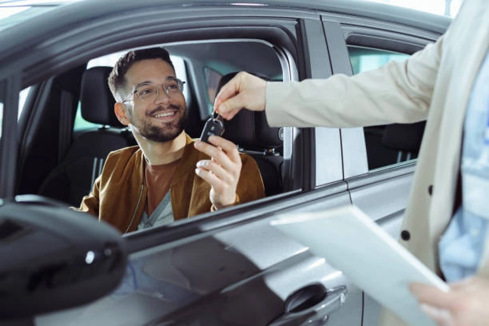 Condutor sorridente está dentro de seu carro e recebe chaves de um corretor que está com papéis nas mãos. Falando sobre se consórcio de carro vale a pena.