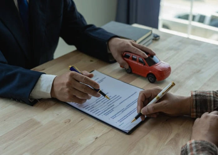 Duas pessoas assinam papel que está sobre um birô. Um dos homens segura uma miniatura de carro vermelho. Explicando quanto custa um seguro de carro por mês.