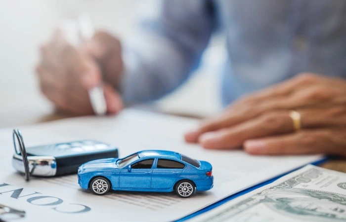 Pessoa assina documento. Sobre a mesa há uma miniatura de carro azul e uma chave de carro. Falando sobre consórcio carro plano de 100 meses.