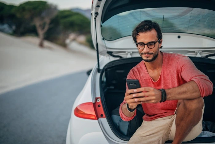 Homem observa dados do seu seguro no smartphone enquanto está sentado na mala do seu carro. Explicando como cotar seguro auto online.