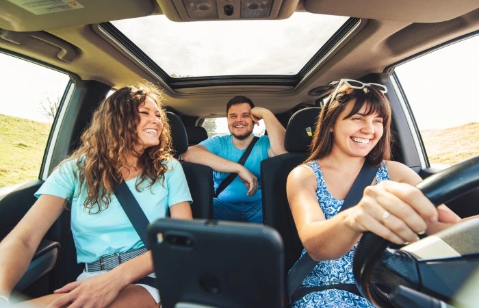 Duas mulheres e um homem sorriem enquanto viajam em um carro. Explicando quanto custa um seguro de carro por mês.