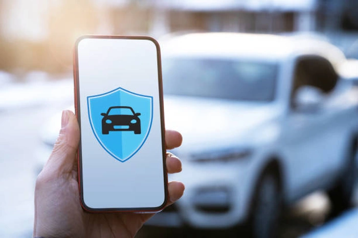 Pessoa segura smartphone com símbolo de proteção auto na tela. Ao fundo há um veículo. Falando sobre como fazer seguro auto online.