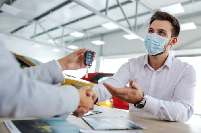 Dois homens dão as mãos. Um entrega as chaves de um carro a outro. O que recebe as chaves está de máscara. Explicando como fazer um consórcio de carros usados.