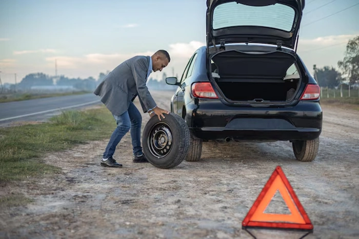Homem prepara troca de pneu de carro. Ele já sinalizou a via com um triângulo. Explicando se seguro auto com assistência 24h vale a pena.